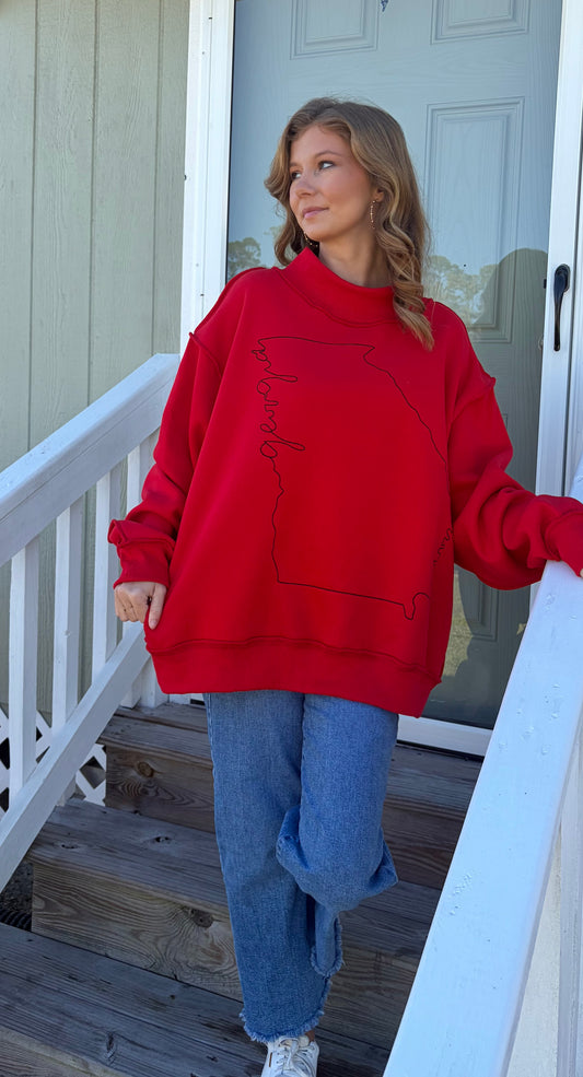 A woman wearing a bright red sweatshirt with a black outline graphic of the state of Georgia and the word 'Georgia' written inside, paired with blue jeans, standing on a wooden porch staircase.