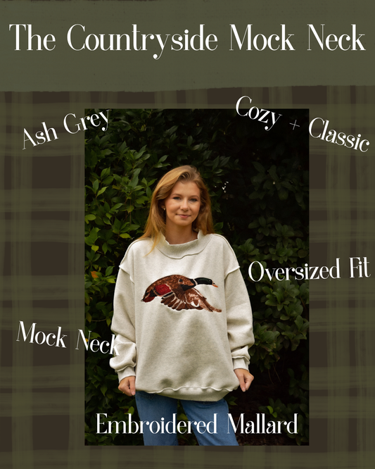 A woman wearing an oversized ash grey mock neck sweatshirt with an embroidered mallard duck graphic on the front, set against a dark green hedge. The image features text overlays describing the product features: 'The Countryside Mock Neck', 'Ash Grey', 'Cozy + Classic', 'Oversized Fit', and 'Embroidered Mallard'.