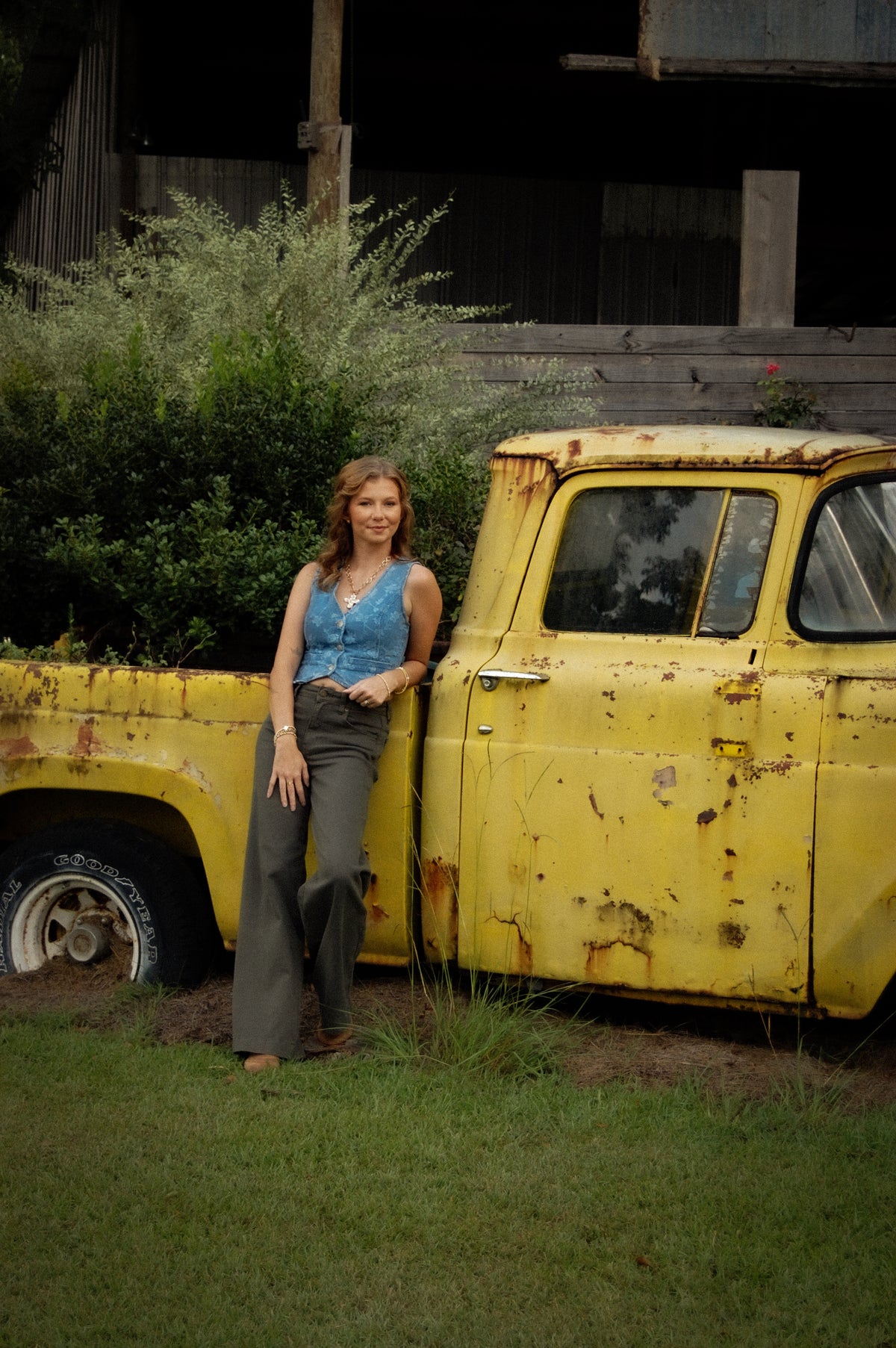 A woman stands outdoors leaning against a weathered yellow pickup truck beside a grassy lawn and rustic building. She is wearing a sleeveless blue denim jacquard vest and dark wide-leg pants.