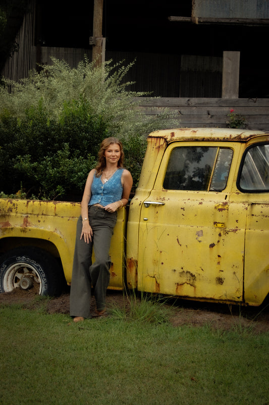 A woman stands outdoors leaning against a weathered yellow pickup truck beside a grassy lawn and rustic building. She is wearing a sleeveless blue denim jacquard vest and dark wide-leg pants.