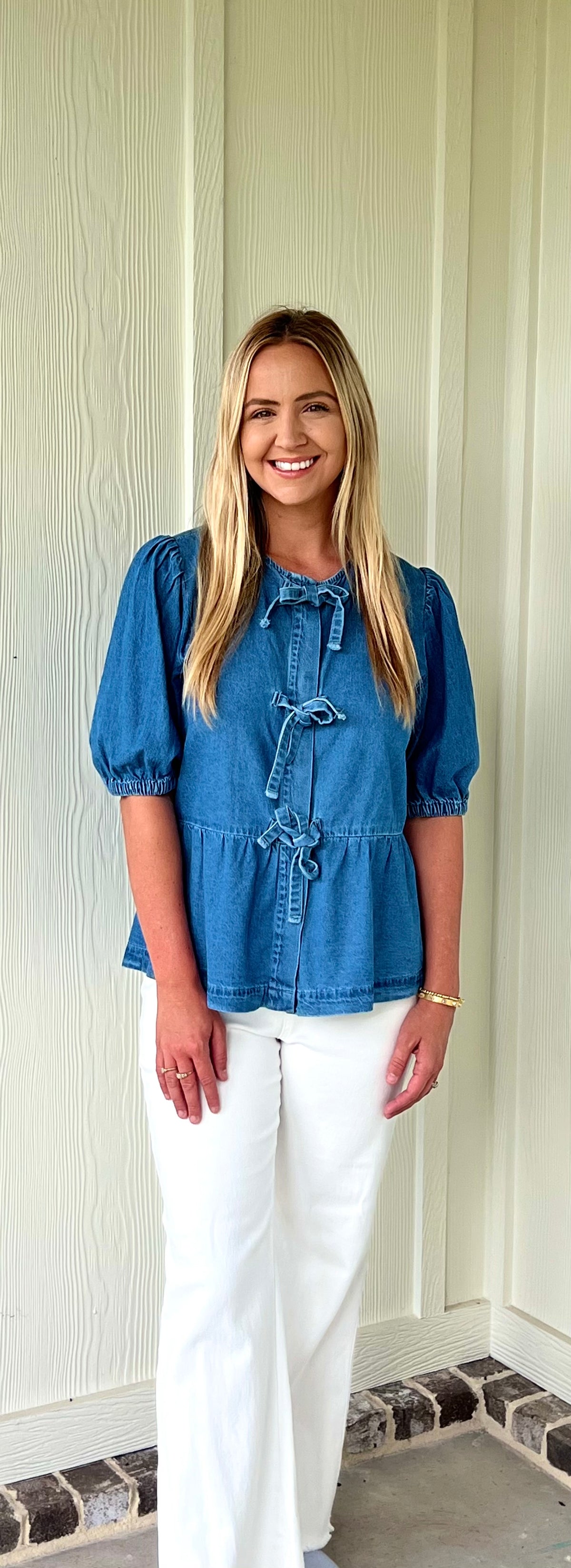 Woman wearing a blue blouse with bow detailing and white pants standing against a white paneled wall.