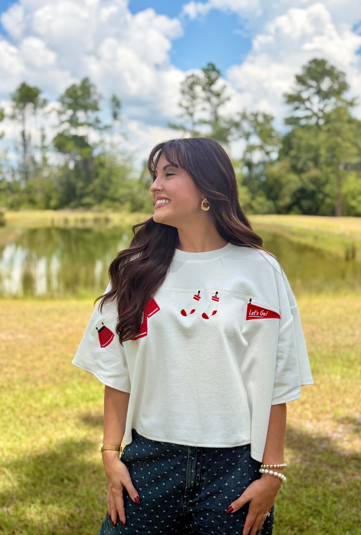 A woman wearing a white short-sleeve graphic t-shirt with red details, standing outdoors in a grassy park with a pond and trees in the background.