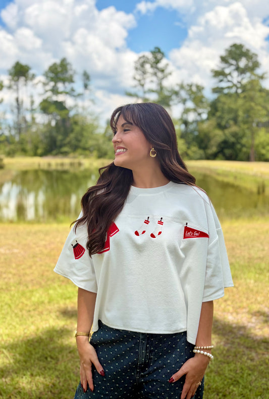 A woman wearing a white short-sleeve graphic t-shirt with red details, standing outdoors in a grassy park with a pond and trees in the background.