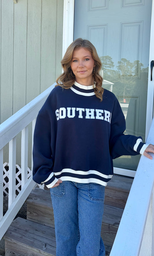 A woman wearing a navy blue and white 'SOUTHERN' varsity sweatshirt and blue jeans, standing on wooden steps in front of a house door.