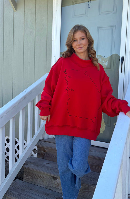 A woman wearing a red oversized sweatshirt with a black outline graphic of the state of Georgia on the front, paired with blue jeans, standing on a wooden porch staircase.