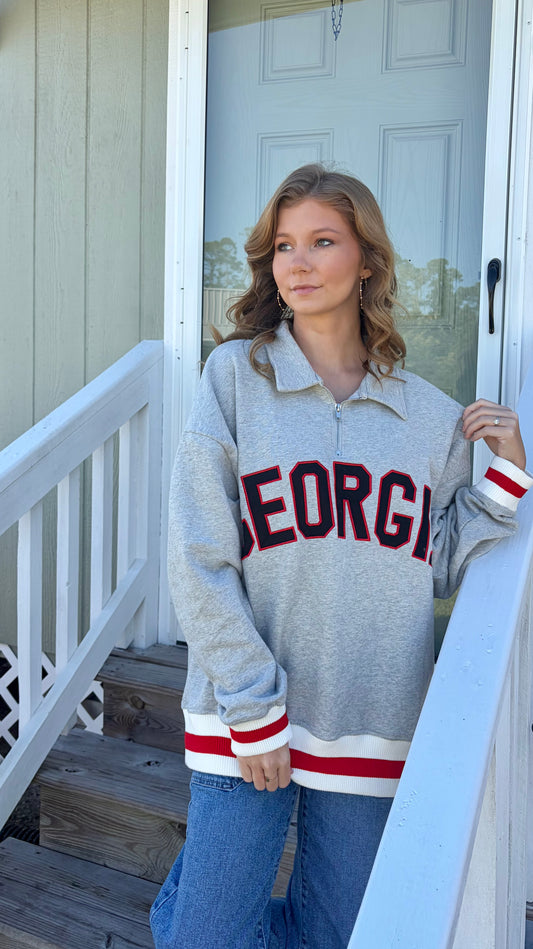 A woman stands on a porch wearing a heather gray quarter-zip sweatshirt with 'GEORGIA' printed across the chest in red and black, paired with blue jeans.