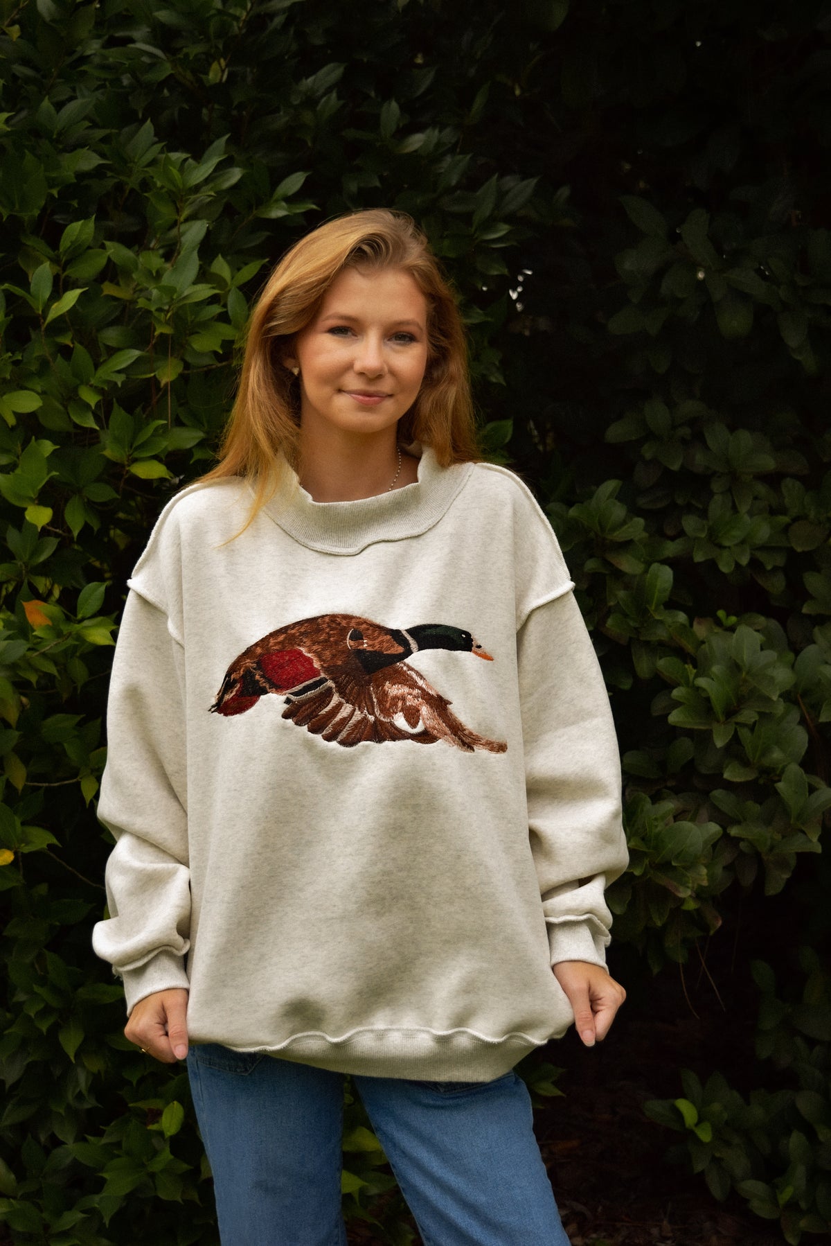 A woman wearing a cream-colored crewneck sweatshirt with a large embroidered duck graphic on the front, paired with blue jeans, standing in front of a dark green leafy background.