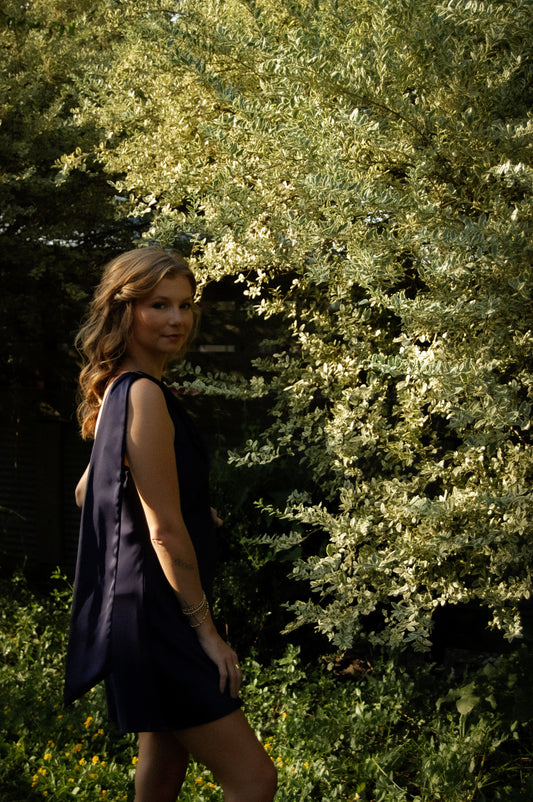 A woman wearing a sleeveless dark navy dress stands outdoors in front of dense green foliage. The dress is open-back and has flowing shoulder details.