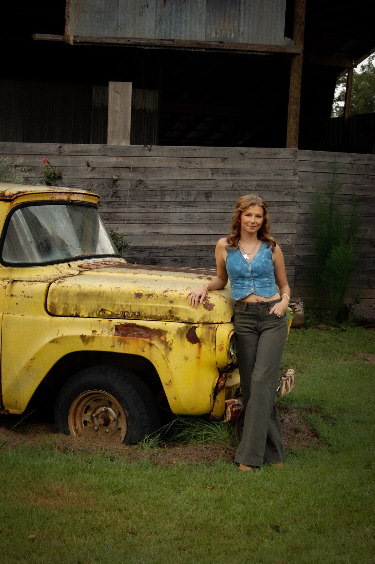 A woman stands outdoors next to a weathered yellow vintage pickup truck in front of a wooden barn wall. She is wearing a denim sleeveless crop top and dark green flared pants.