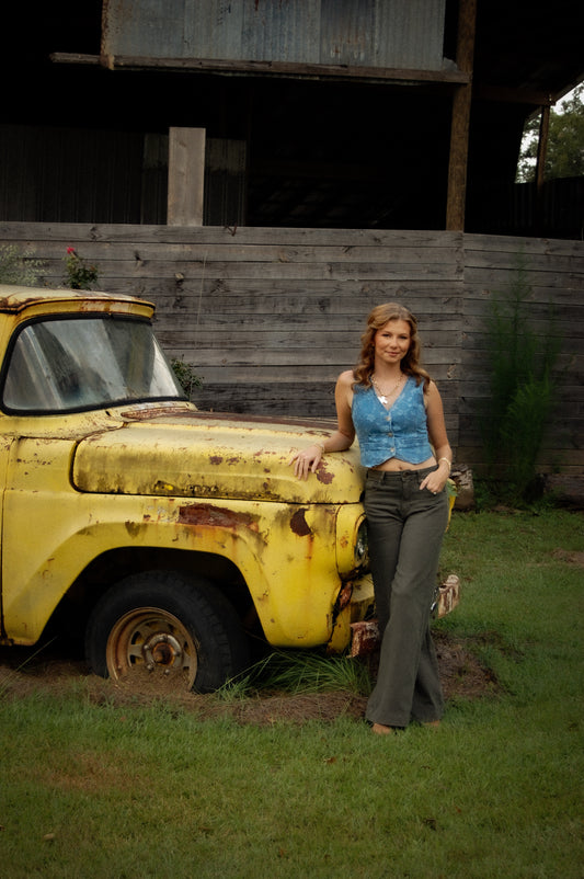A woman stands outdoors next to a weathered yellow vintage pickup truck in front of a wooden barn wall. She is wearing a denim sleeveless crop top and dark green flared pants.