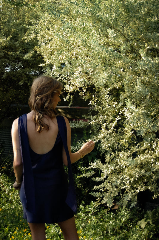 A woman wearing a navy blue sleeveless dress stands outdoors in a garden, seen from behind as she looks at green foliage. The deep navy dress features an open back, as well as an elegant shoulder drape.