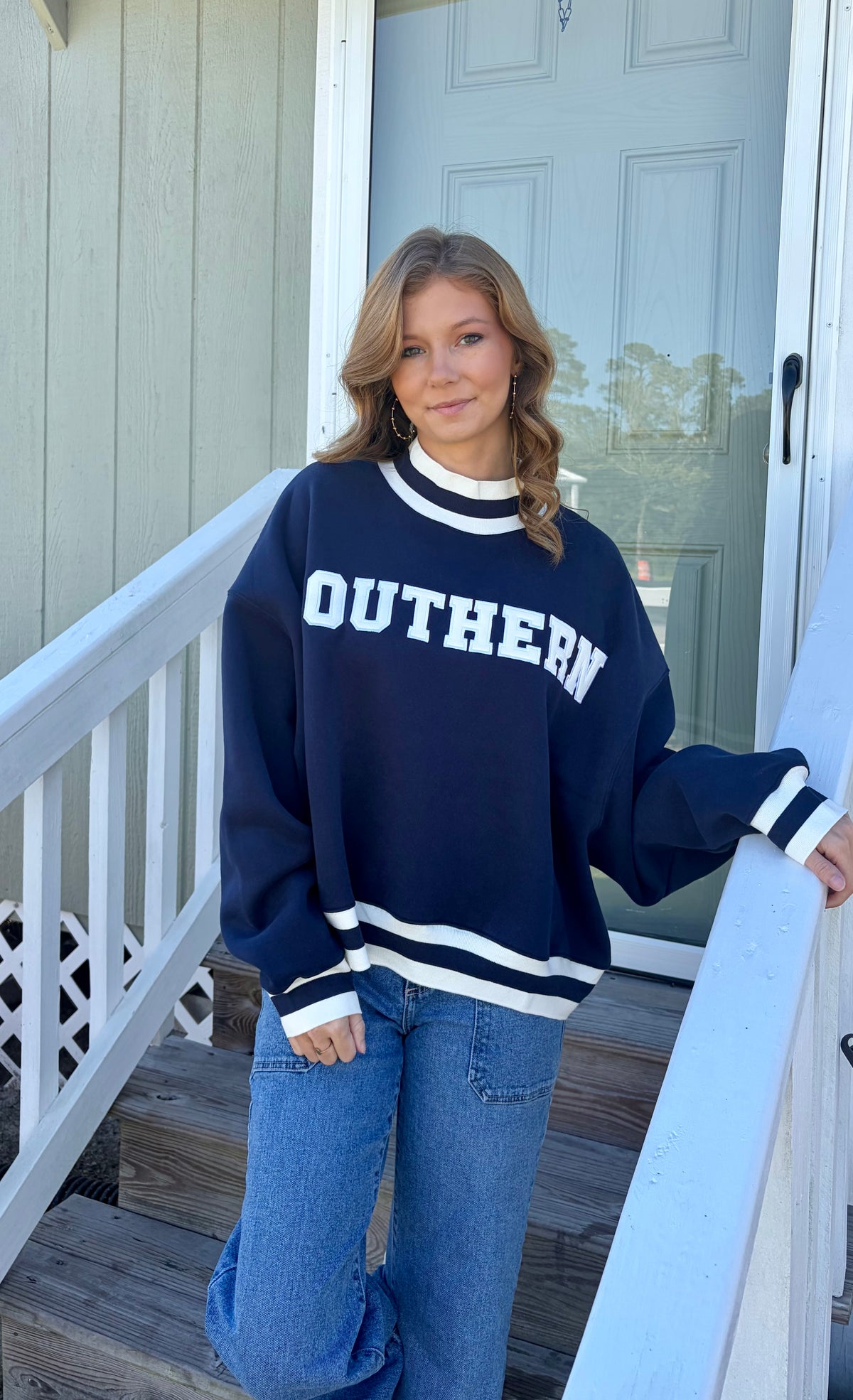 A woman stands on outdoor wooden steps wearing a navy blue crewneck sweatshirt with 'SOUTHERN' printed in white across the chest, paired with blue jeans.