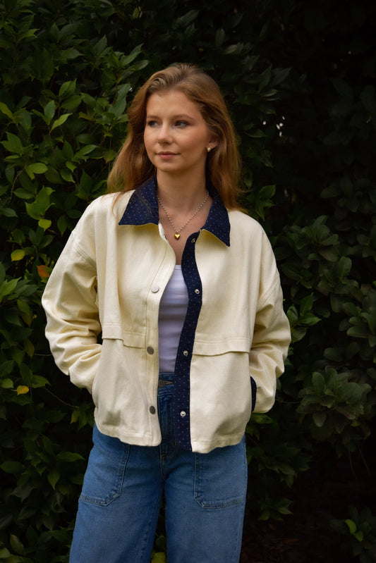A woman wearing a cream-colored jacket with a white polka dot and dark navy blue collar and front placket, layered over a white top and blue jeans, standing in front of a dark green leafy background.