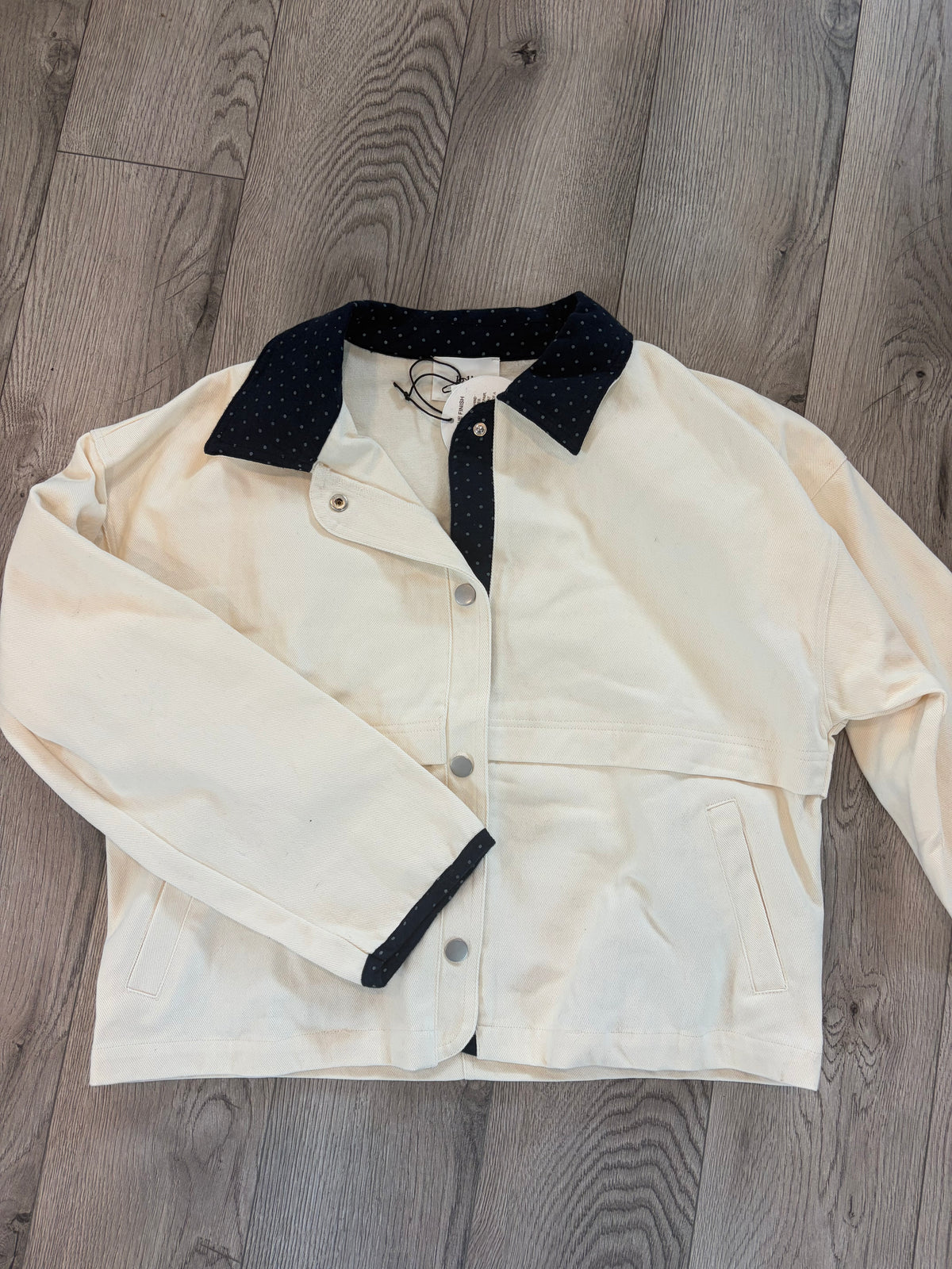 An off-white utility jacket with a contrasting navy polka-dot collar and navy cuffs, laid flat on a gray wood plank floor.