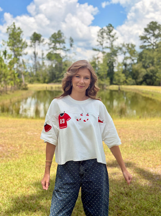 A woman stands outdoors in a grassy field with a pond and trees in the background, wearing a white cropped t-shirt with red and black graphic details on the front and sleeves, paired with dark blue polka-dot pants.