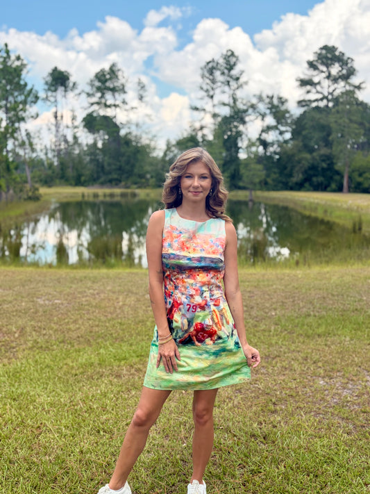 A woman stands outdoors in a grassy field with a pond and trees in the background, wearing a sleeveless mini dress with a colorful abstract floral print and white sneakers.